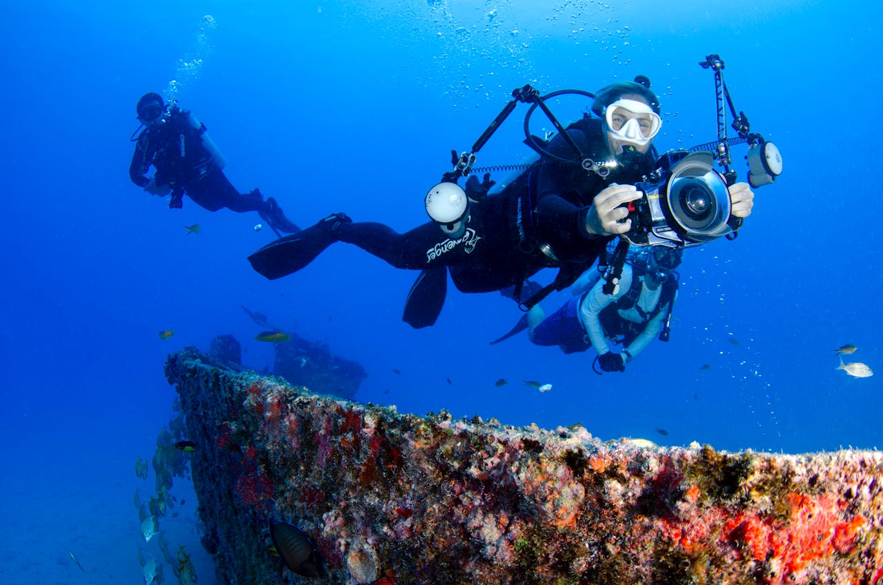 Home Two scuba divers capturing the vibrant marine life near an underwater wreck in crystal clear waters.